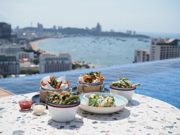 Rooftop dining setup overlooking Pattaya Bay with an infinity pool, gourmet Asian fusion dishes, and a panoramic skyline and sea view in the background.