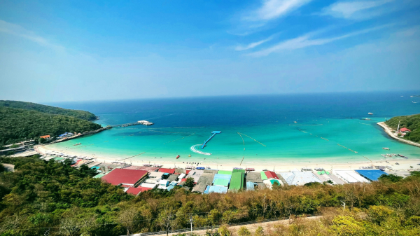 A panoramic aerial view of Koh Larn (Coral Island) near Pattaya, showing turquoise waters, white sand beaches, water activities, and colorful beachfront buildings surrounded by lush hills.